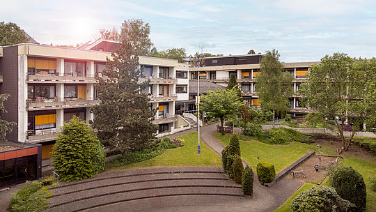 Mehrstöckige Wohngebäude mit Balkonen, umgeben von Bäumen und einem gepflasterten Innenhof mit Sitzstufen.