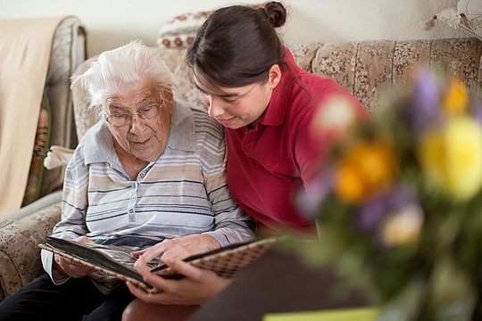 Zwei Personen sitzen auf einem Sofa, eine hält ein Fotoalbum, die andere lehnt sich zum Betrachten vor.