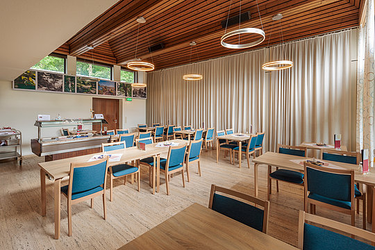 Speisesaal mit mehreren Holztischen und blauen Stühlen, Decke mit Holzverkleidung und runden Deckenleuchten, große Fenster mit hellen Vorhängen.