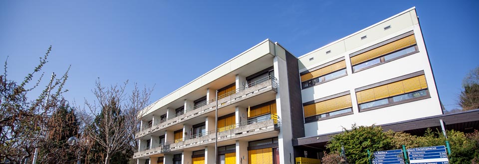 Mehrstöckiges Gebäude mit Balkonen und gelben Sonnenschutzmarkisen, umgeben von Bäumen und Sträuchern