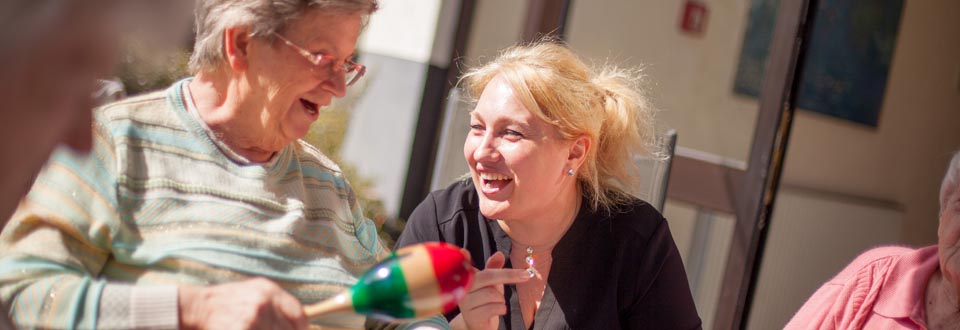Drei Personen sitzen an einem Tisch, eine hält ein buntes Rasselinstrument, eine weitere Person ist mit einer Handbewegung beteiligt