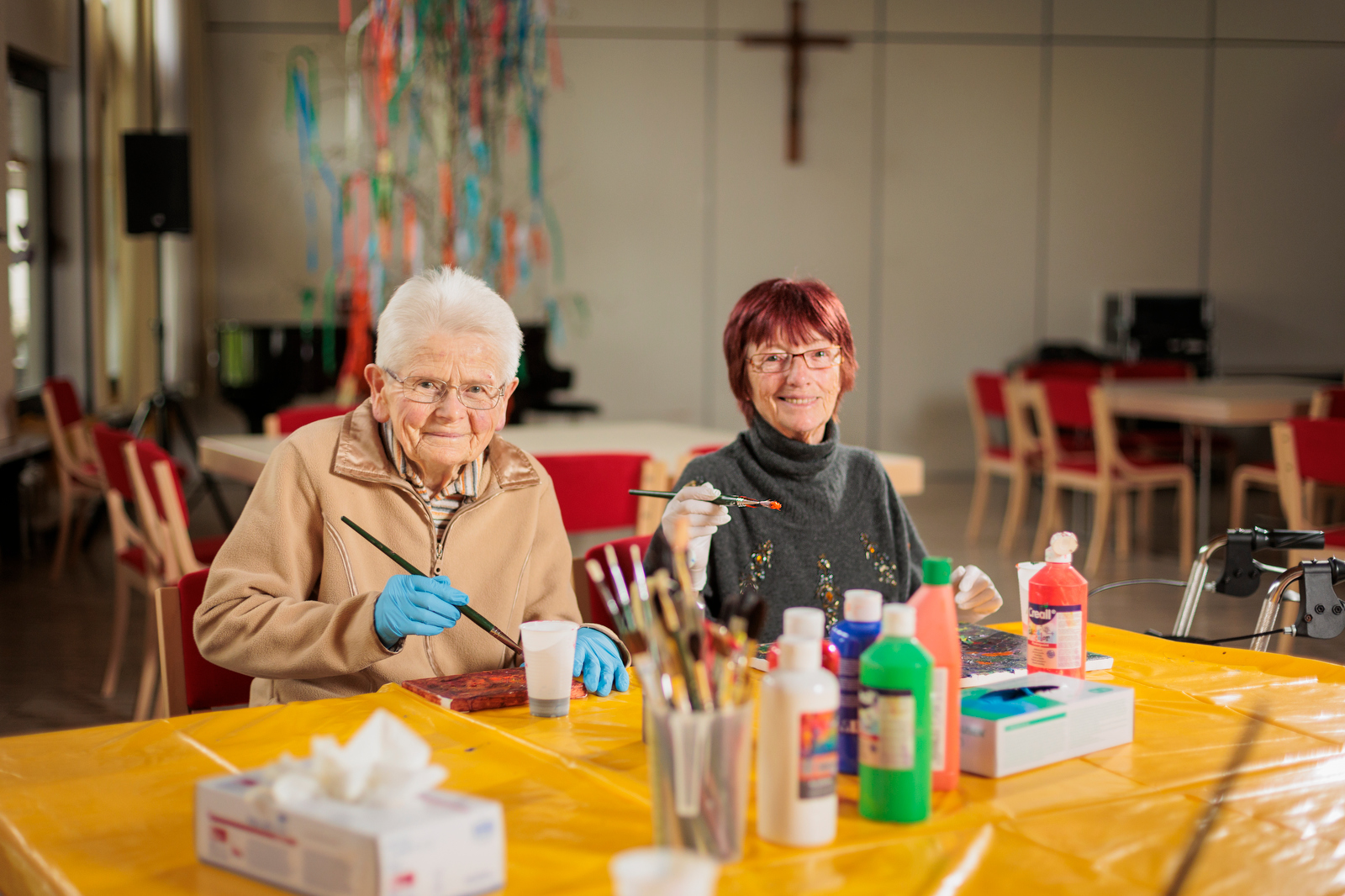 Zwei Personen sitzen an einem Tisch mit Malutensilien und bunten Farben in einem Raum mit Kreuz an der Wand.